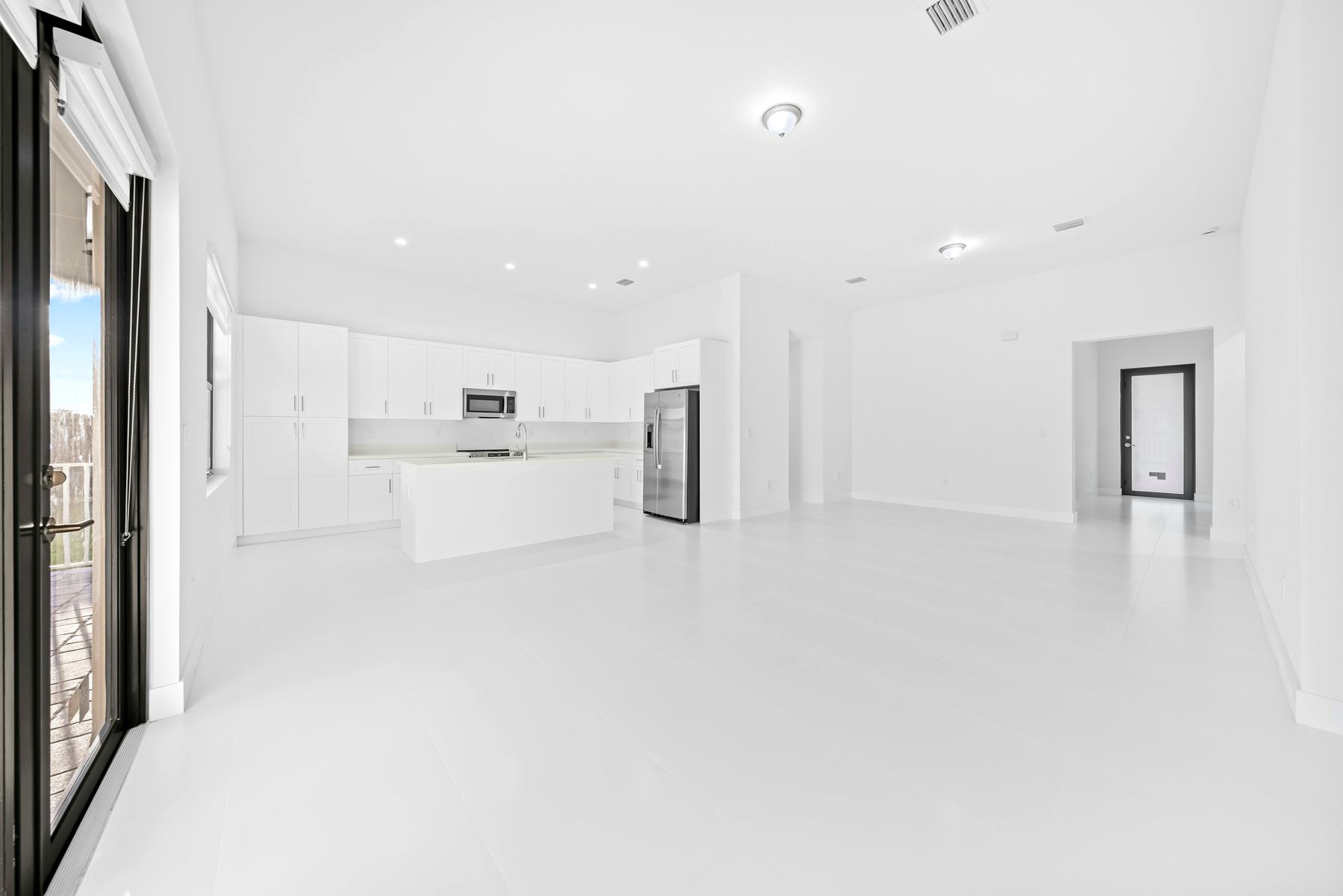 A bright, empty white kitchen and living area with daylight coming through a glass door.